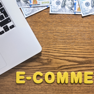 Laptop sobre mesa de madeira escura com notas de dólar espalhadas e o texto "E-COMMERCE" em letras amarelas no canto inferior.