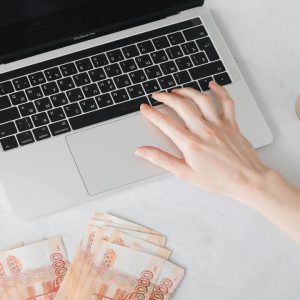 Mão sobre um laptop com notas de Real espalhadas e um copo de café, simbolizando ganho de dinheiro na internet com PLR.