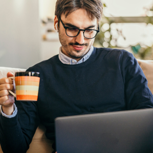 Homem de óculos tomando café e trabalhando em um laptop no sofá, avaliando os prós e contras do mercado PLR.