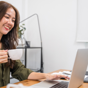 Mulher sorrindo, tomando café e trabalhando em um laptop, em um escritório moderno, representando o sucesso com nichos de PLR.